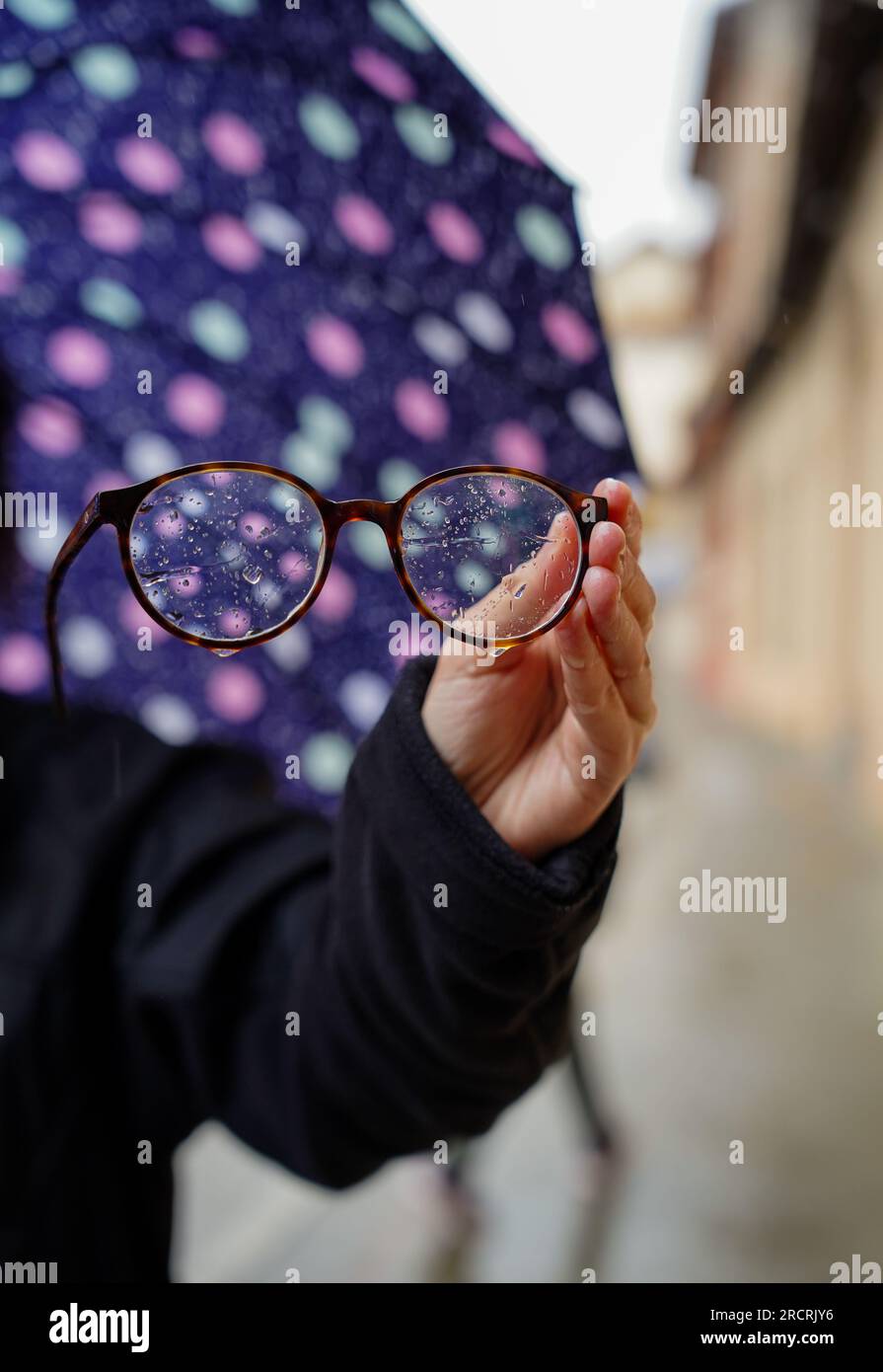 Eine Hand, die eine nasse Brille mit Wassertropfen und einen Regenschirm im Hintergrund hält Stockfoto