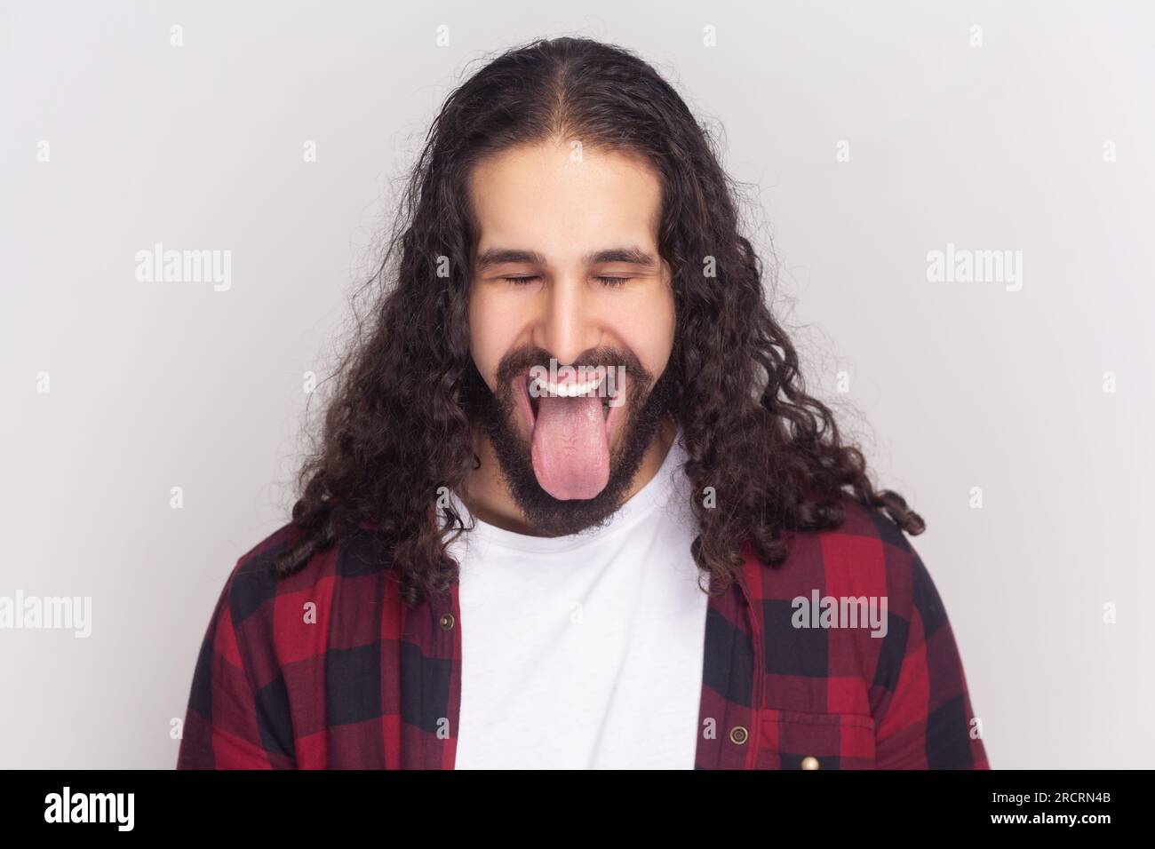 Ein komischer bärtiger Mann im karierten roten Hemd sticht aus der Zunge heraus, macht fröhliche Grimace, drückt positive Emotionen aus, töricht mit Freunden. Studioaufnahmen im Innenbereich isoliert auf grauem Hintergrund. Stockfoto