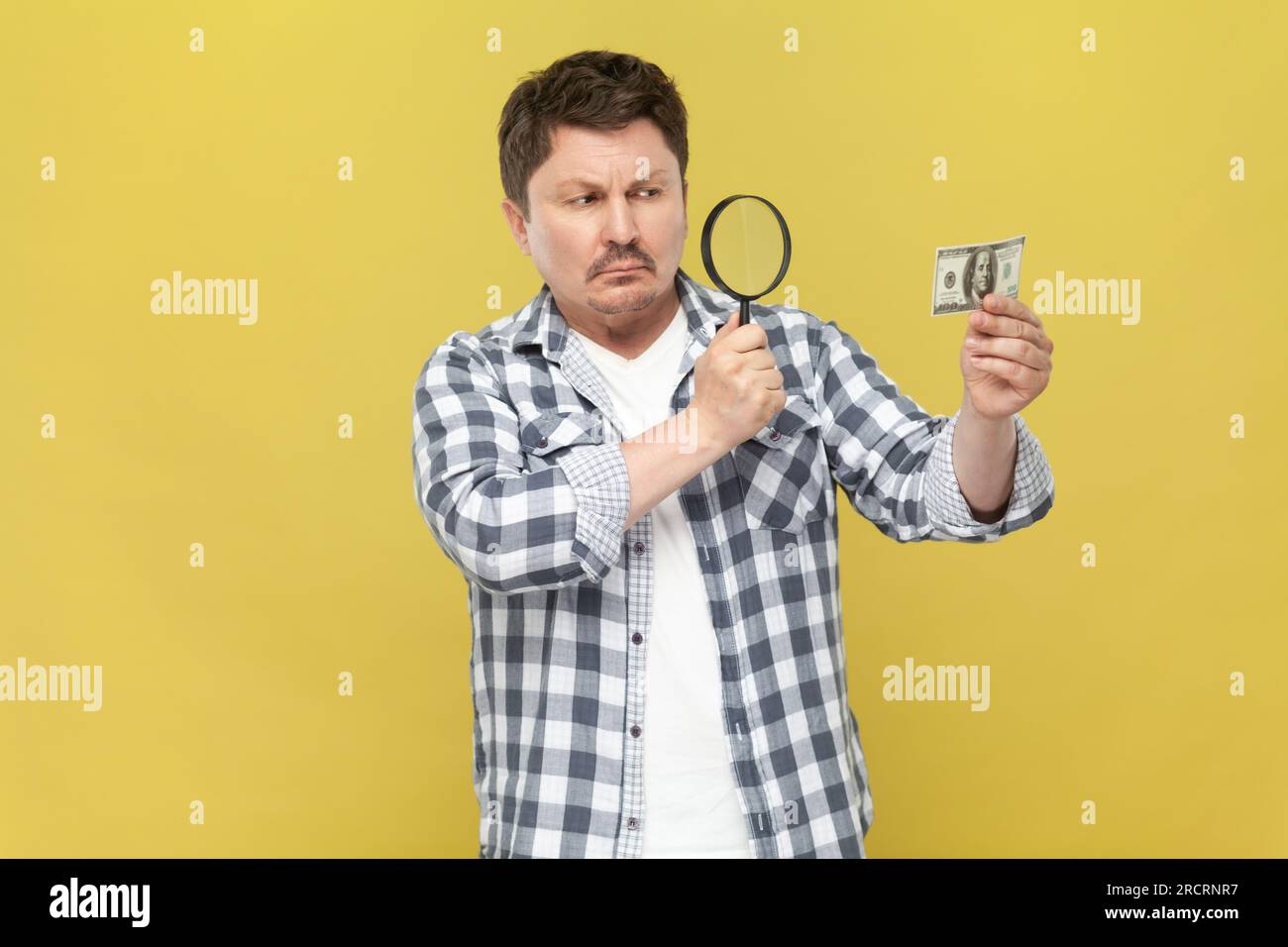 Porträt eines ernsthaften konzentrierten Mannes mittleren Alters in einem lässigen karierten Hemd, der durch die Lupe in den Dollar schaut und die Echtheit der Banknote prüft. Innenstudio-Aufnahme isoliert auf gelbem Hintergrund. Stockfoto