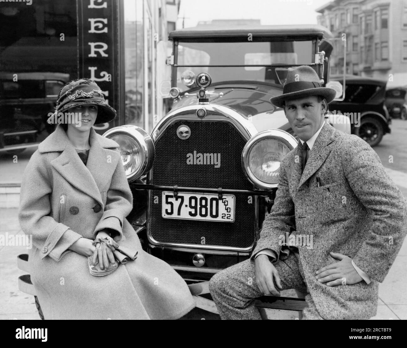 San Francisco, Kalifornien: ca. 1922 Ein Paar vor dem Peerless-Autohaus sitzt auf der Stoßstange ihres 1922 Peerless mit Colorado-Kennzeichen. Stockfoto
