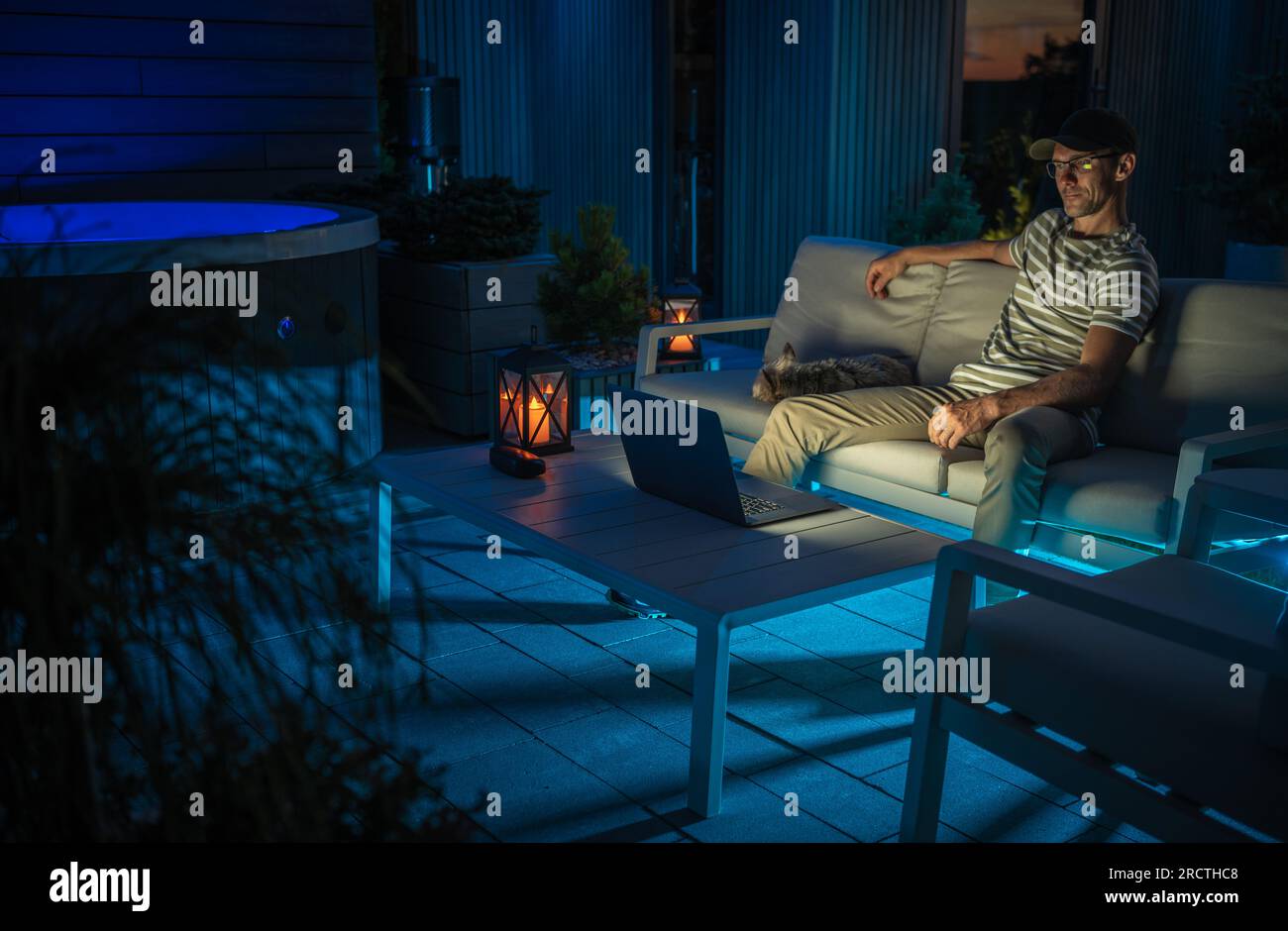 Der Weiße Mann in seinem 40s. Jahrhundert sieht sich Filme auf seinem Laptop in seinem modernen, blau beleuchteten Garten an Stockfoto