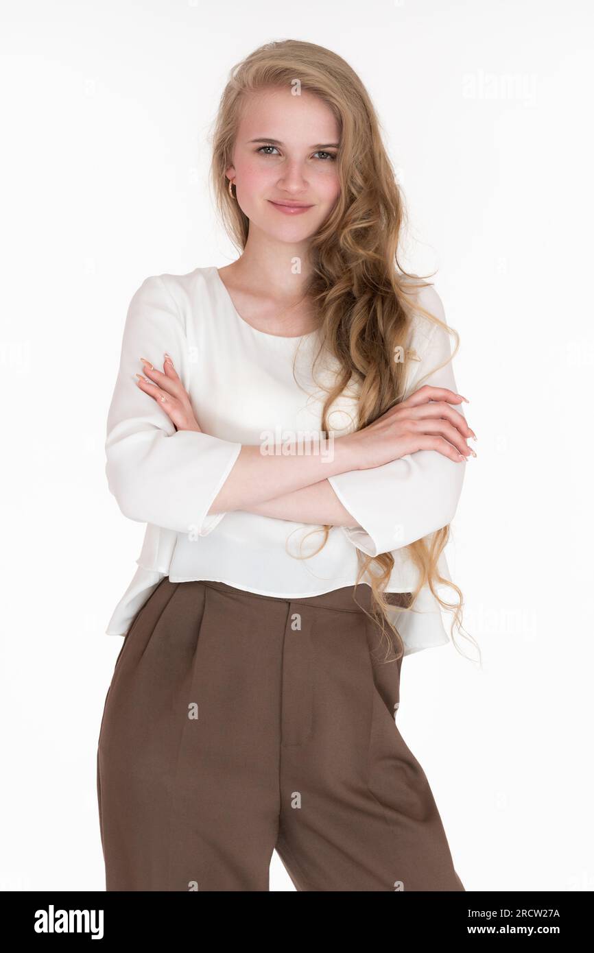 Lächelnde junge blonde Frau mit gekreuzten Armen, die in die Kamera schaut. Herrisch-junge Geschäftsfrau, 21 Jahre alt, in weißer Bluse und brauner Hose Stockfoto