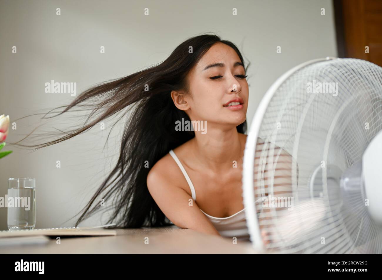 Eine wunderschöne Asiatin, die sich überhitzt fühlt, kühlt sich mit einem elektrischen Ventilator ab, während sie sich an einem heißen Sommertag in ihrem Wohnzimmer ausruht. Stockfoto