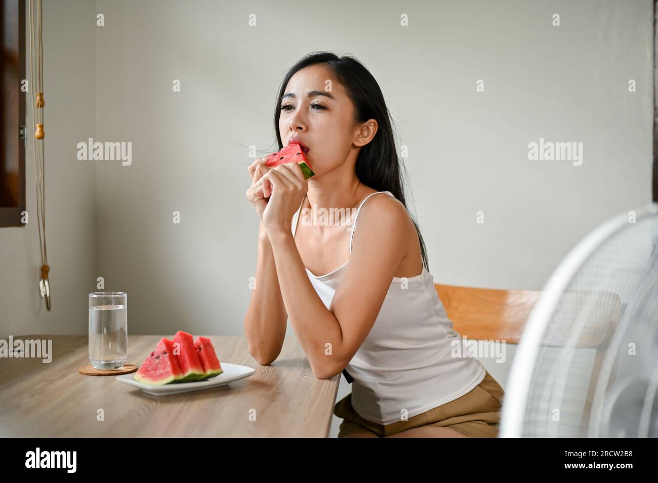 Eine wunderschöne asiatische Frau, die sich durch das heiße Wetter unwohl fühlt, isst Wassermelone und sitzt vor einem elektrischen Ventilator, um sich zu Hause zu erfrischen Stockfoto