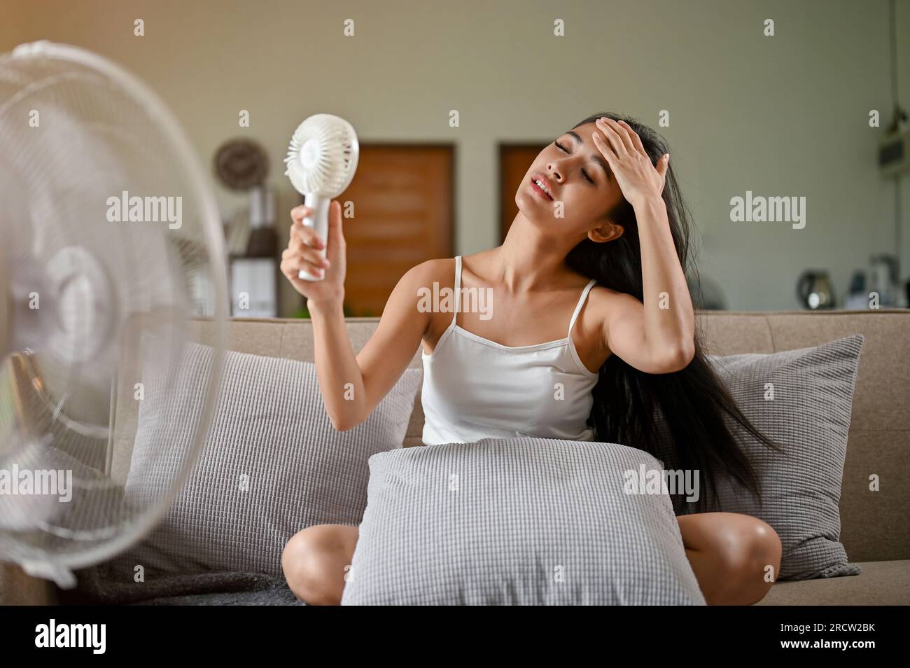 Eine wunderschöne Asiatin, die sich überhitzt und müde fühlt und unter einem Hitzeanfall leidet, erfrischt sich mit einem handlichen Ventilator und einem elektrischen Ventilator Stockfoto