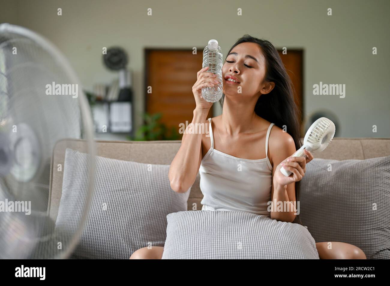 Eine müde und heiße Asiatin versucht sich mit einem elektrischen Ventilator abzukühlen und berührt ihr Gesicht mit einer Kühlwasserflasche, während sie darauf sitzt Stockfoto