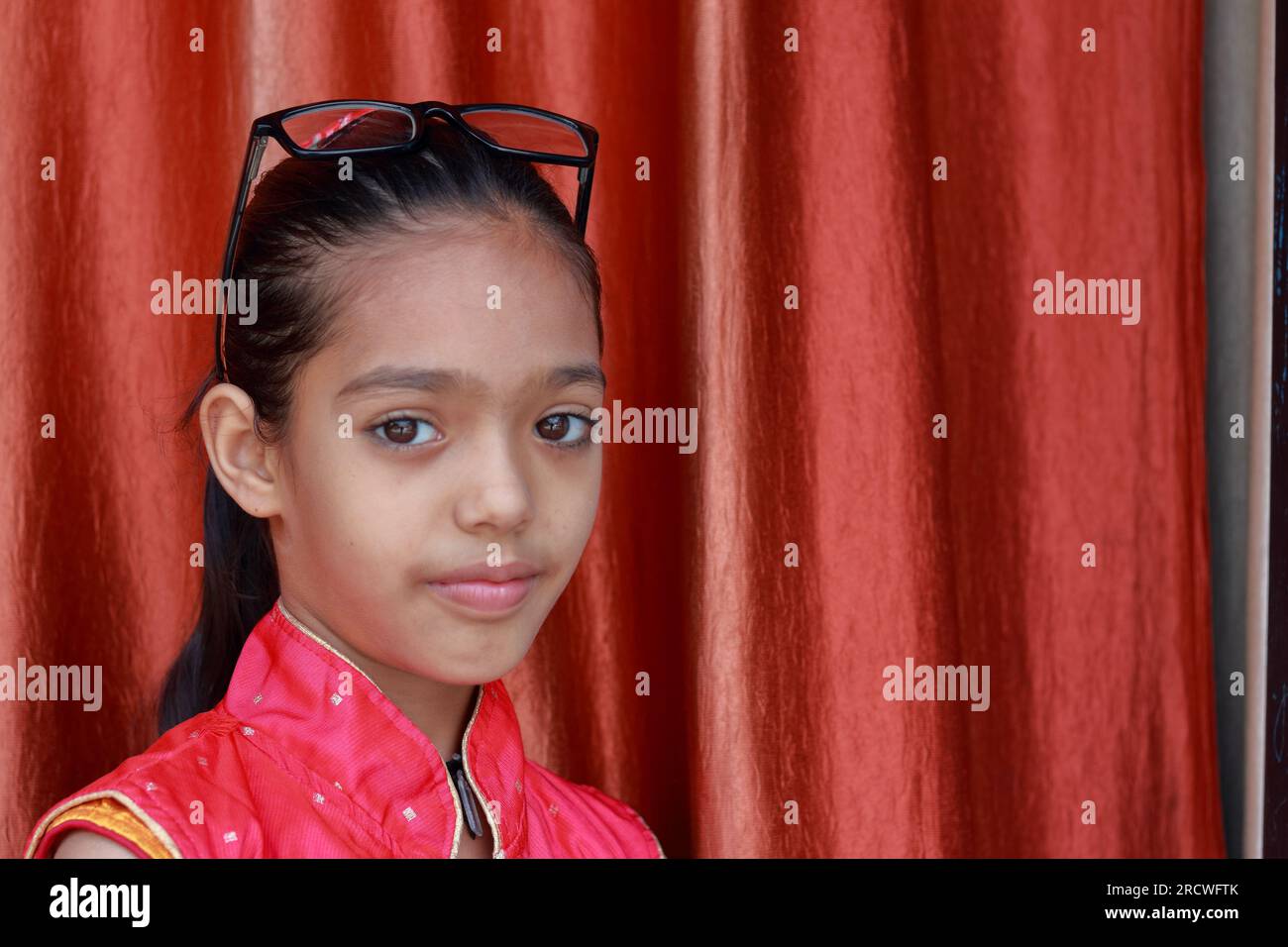 Ein indisches kleines hübsches Mädchen, das mit ihrer Brille in verschiedenen Posen in rotem Kleid rumfummelt. Stockfoto