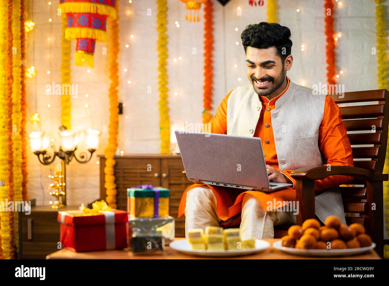 Glücklicher junger indischer Mann, der während der diwali Festival Feier im dekorierten Haus ein Notebook benutzte, vor Geschenken und Geschenken, indem er in die Kamera schaute - Konzept Stockfoto
