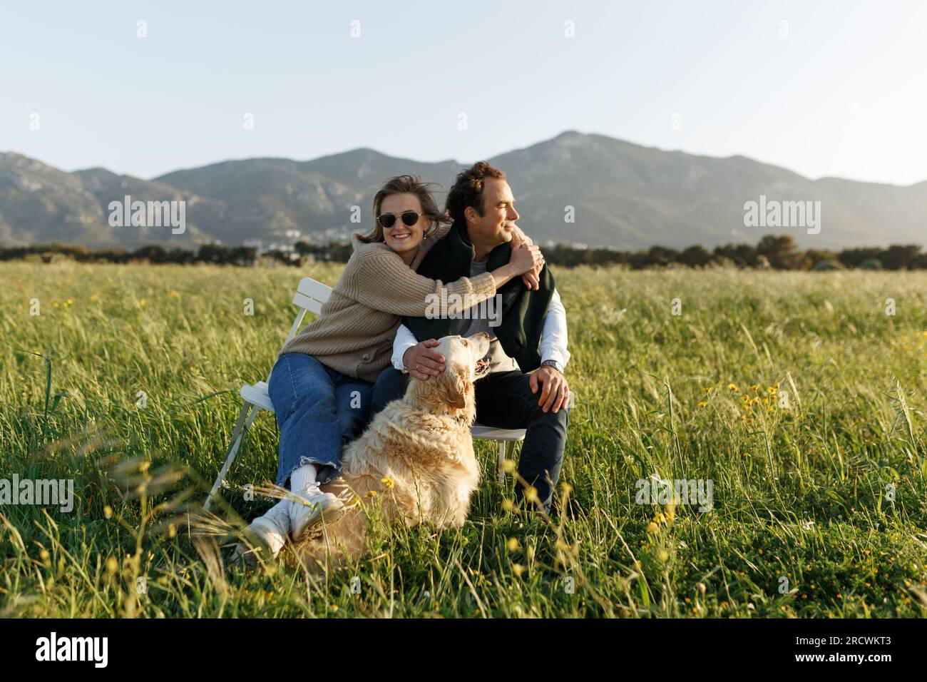 Ein junges glückliches Paar mit Hund, das auf Stühlen im Federrasenfeld am Sommertag bei Sonnenuntergang sitzt. Stockfoto