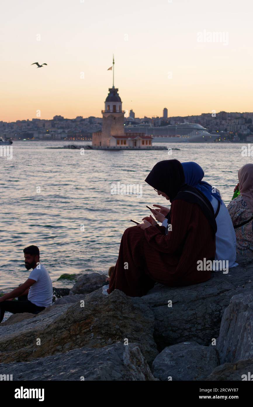 Die Menschen sitzen auf Felsen und genießen den Sonnenuntergang über dem Bosporusmeer und dem Maidens Tower von Uskudar, Istanbul, Türkei. Kreuzfahrtschiff hinten. Stockfoto