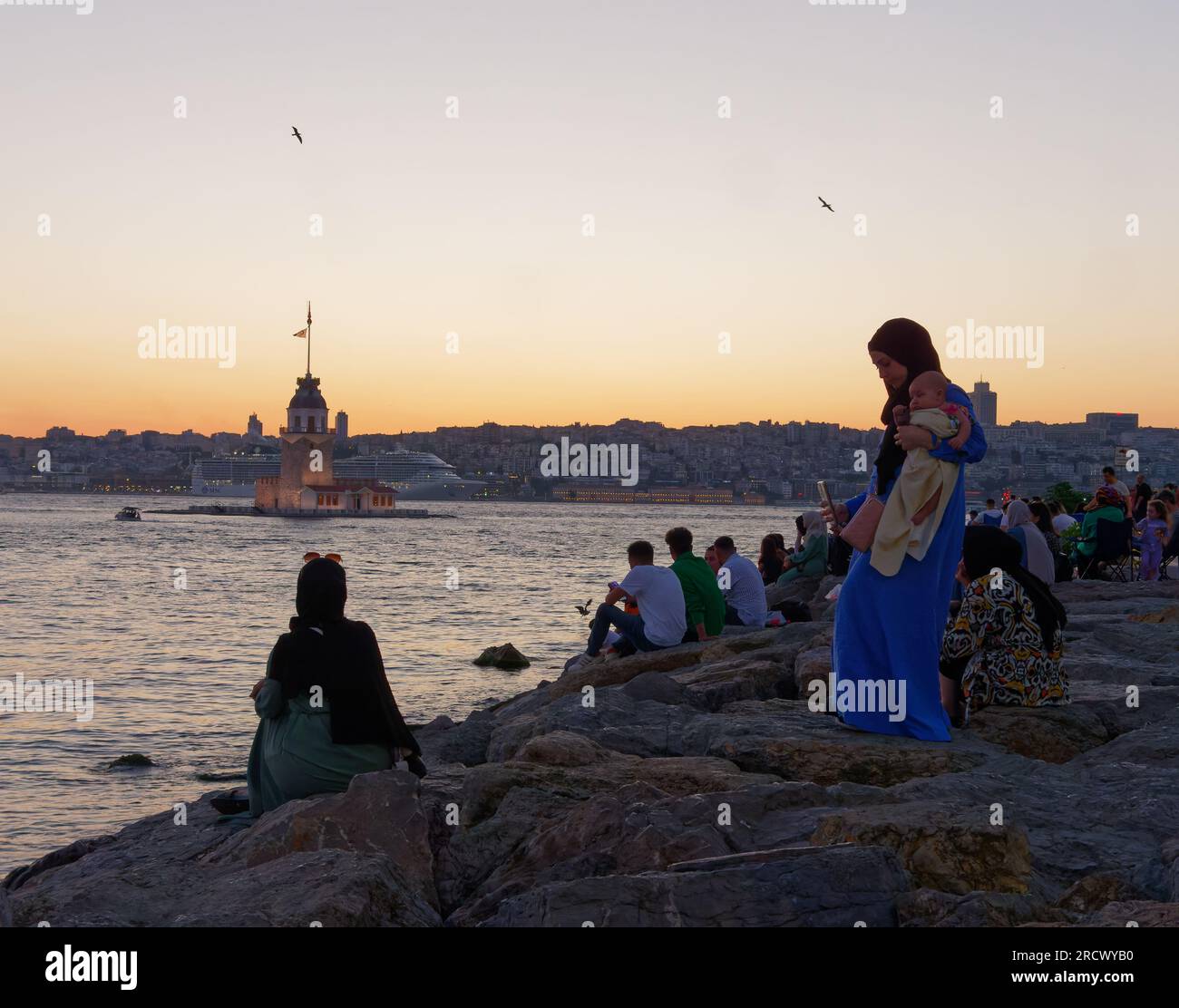 Die Menschen sitzen auf Felsen und genießen den Sonnenuntergang über dem Bosporusmeer und dem Maidens Tower von Uskudar, Istanbul, Türkei. Kreuzfahrtschiff hinten. Stockfoto