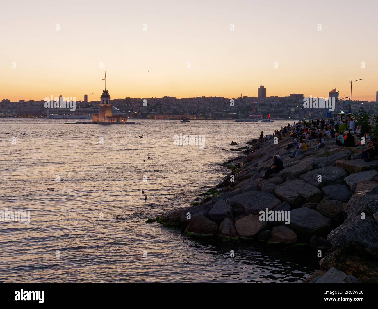 Die Menschen sitzen auf Felsen und genießen den Sonnenuntergang über dem Bosporusmeer und dem Maidens Tower von Uskudar, Istanbul, Türkei. Kreuzfahrtschiff hinten. Stockfoto
