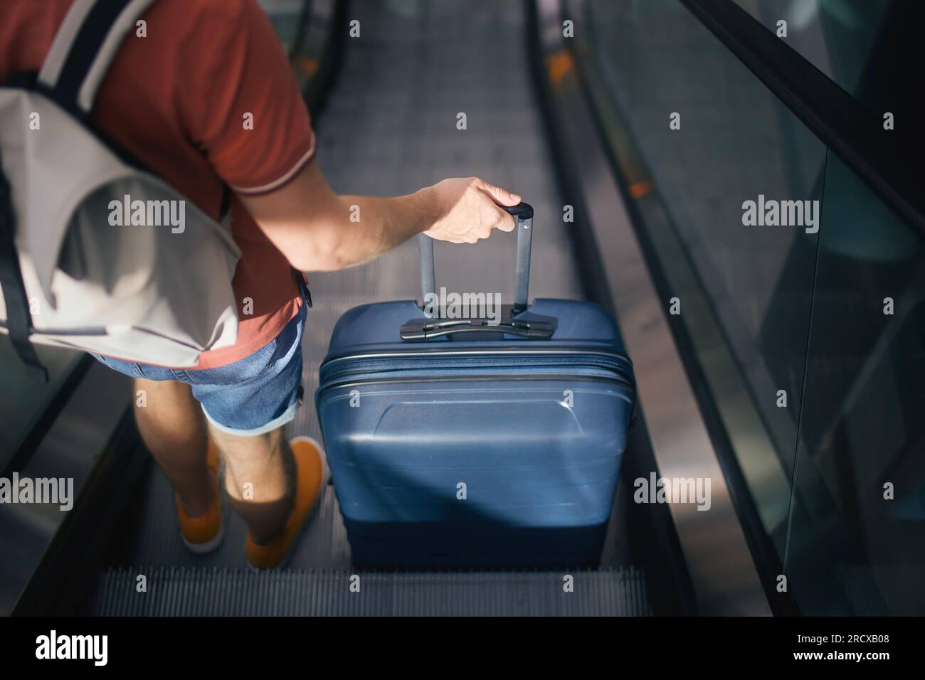Reisende, die durch den Flughafenterminal laufen. Rückansicht eines Mannes mit Koffer und Rucksack, der auf dem Ekalator steht. Der Passagier ist reisebereit. Stockfoto