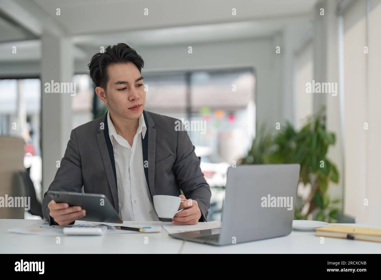 Fokussierter junger Geschäftsmann aus Asien, der am Schreibtisch sitzt und sicherstellt, dass sein Unternehmen genug Geld für die Finanzplanung verdient Stockfoto