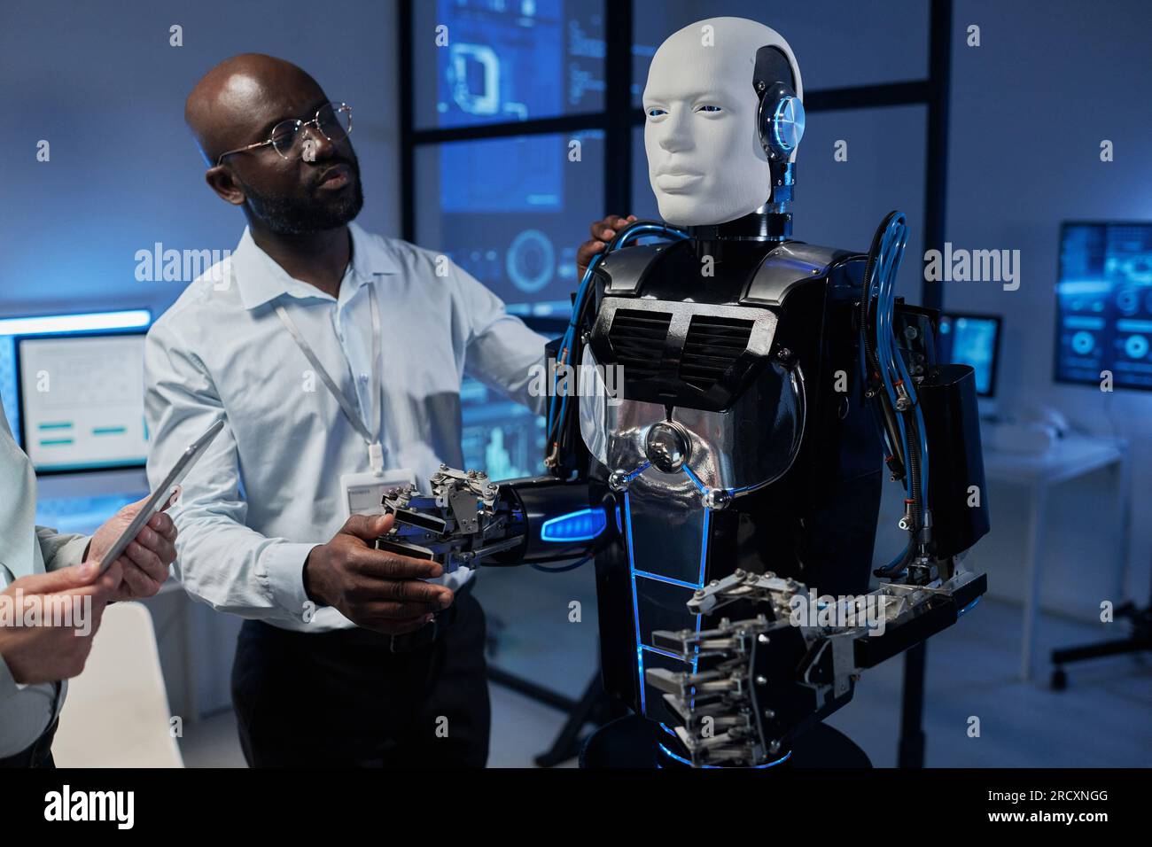 Ingenieur, der den Roboter über ein digitales Tablet steuert, während sein Kollege seinen Roboterarm hält und dabei hilft, sich zu bewegen Stockfoto