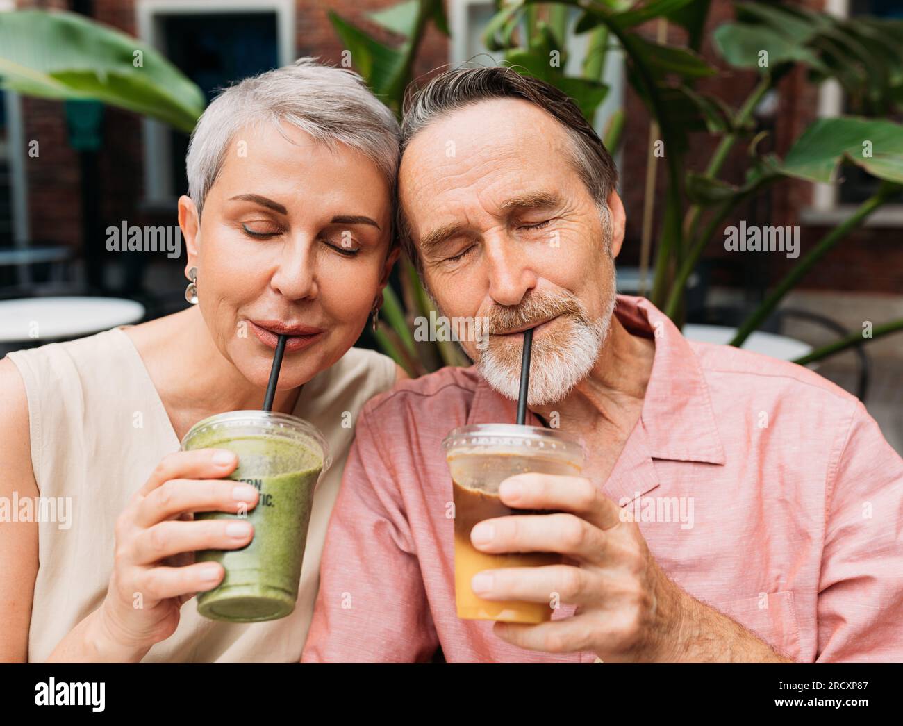 Reifes Paar, das einen Smoothie trinkt. Ehefrau und Ehemann trinken Cocktails, während sie Kopf an Kopf mit geschlossenen Augen sitzen. Stockfoto