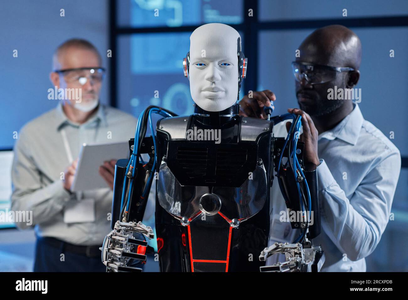 Afroamerikanischer Ingenieur, der Roboter mit Werkzeugen repariert, während sein Kollege ihn im Hintergrund programmiert Stockfoto