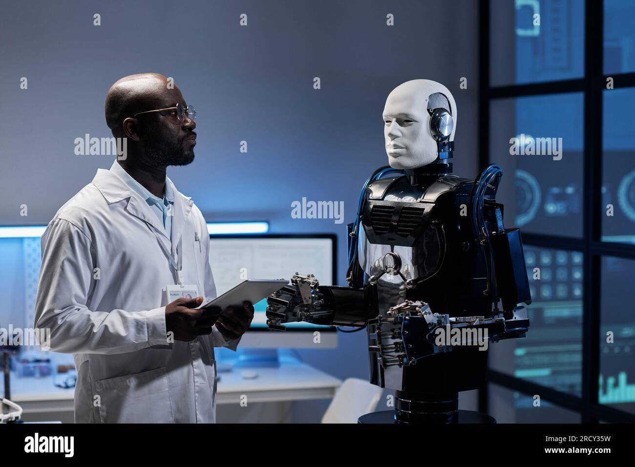 Afroamerikanischer Ingenieur mit weißem Mantel, der an einem Tablet-pc arbeitet, während sich ein Roboter im Büro befindet Stockfoto