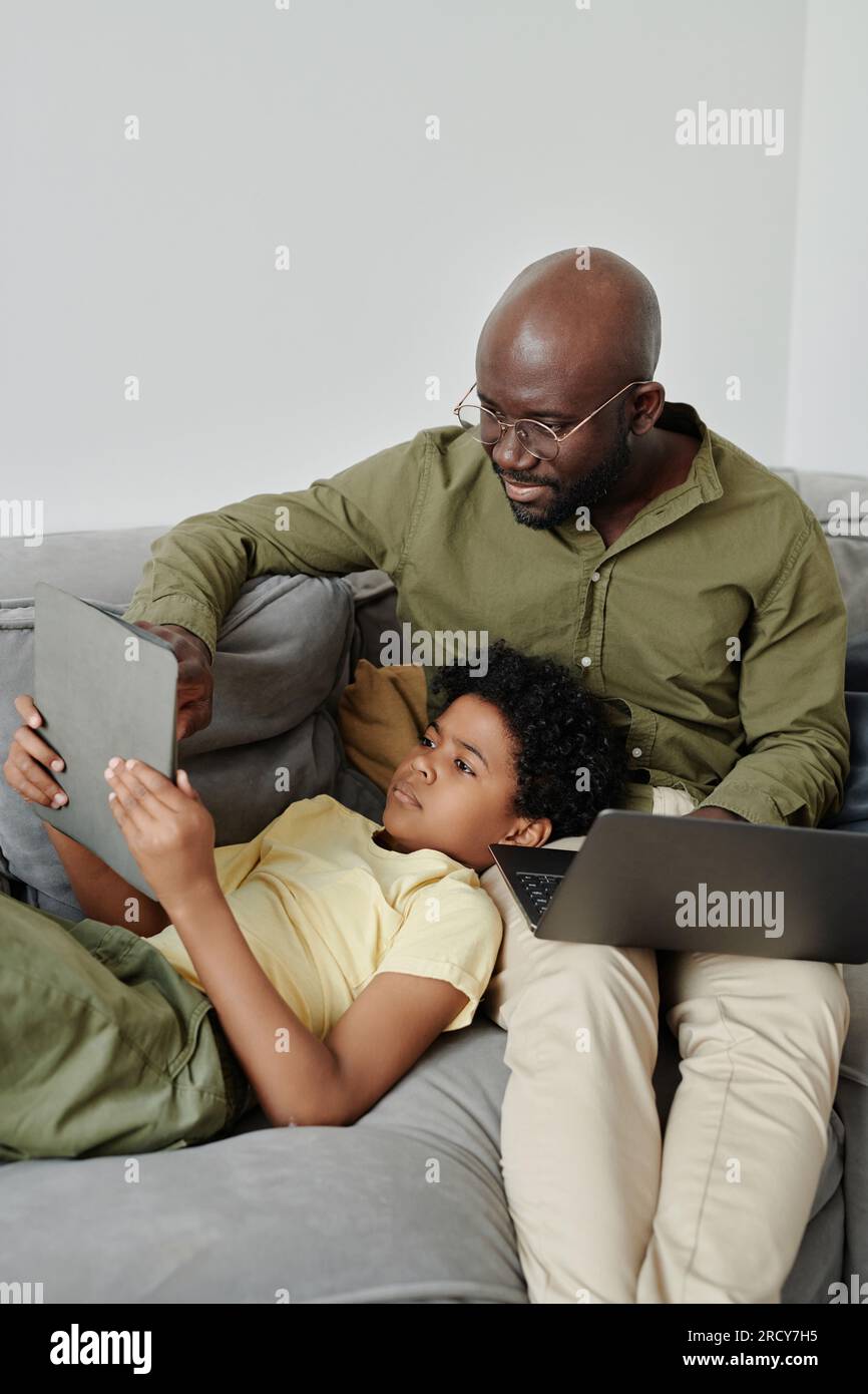 Vertikales Bild eines afroamerikanischen Vaters, der seinem Sohn hilft, einen Tablet-pc zu verwenden, während er auf dem Sofa im Zimmer an einem Laptop arbeitet Stockfoto