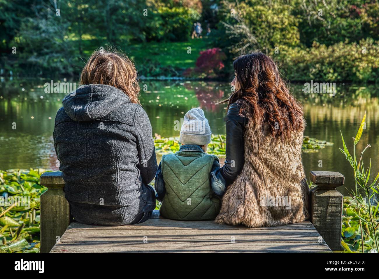 Eine dreiköpfige Familie, die auf einem Steg am See sitzt, mit einem kleinen Kind in der Mitte zwischen mom und Dad. Rückansicht einer Familie mit 1 Kindern, die auf einem Kai sitzt Stockfoto