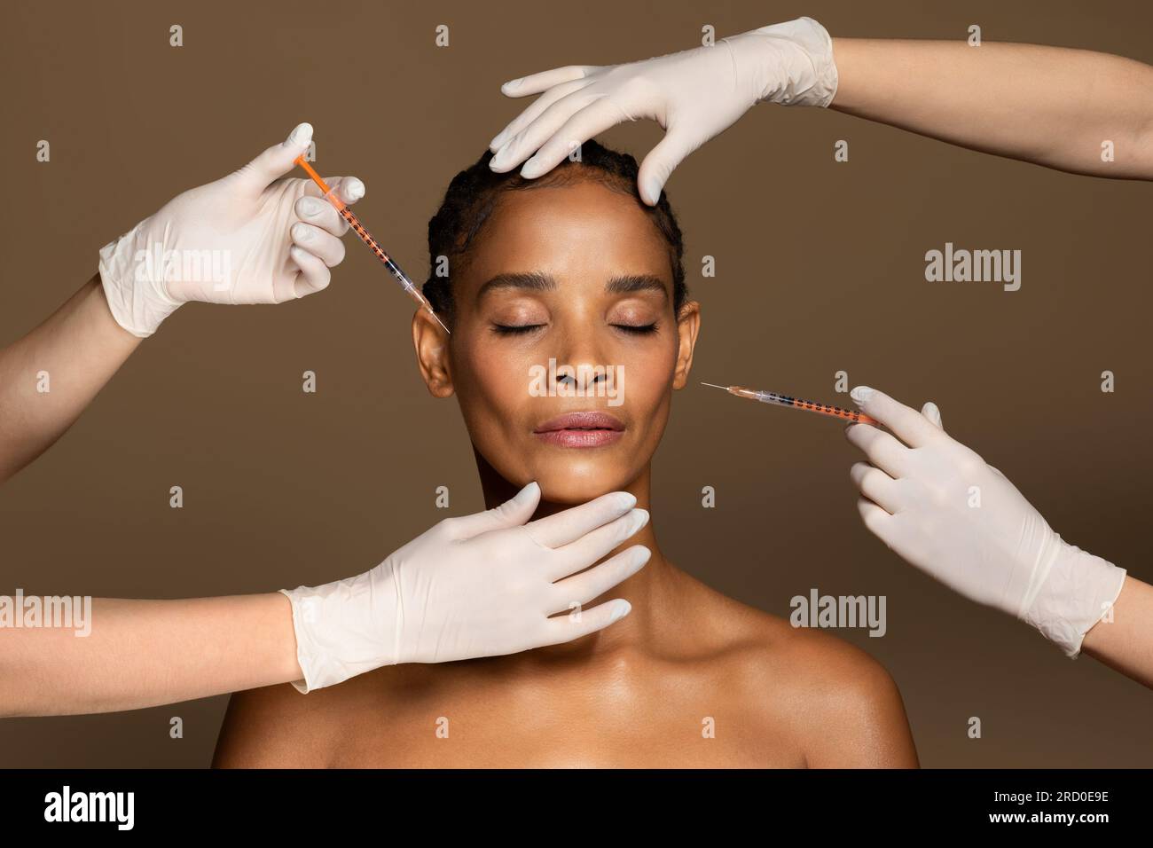 Attraktive afroamerikanische Frau mittleren Alters, die von zwei Kosmetikern Gesichtsspritzen bekommt, brauner Hintergrund Stockfoto