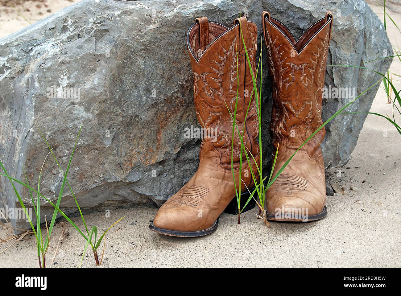 Braune Stiefel im westlichen Stil auf Sand an einem großen Felsen und grünen Grashalmen Stockfoto