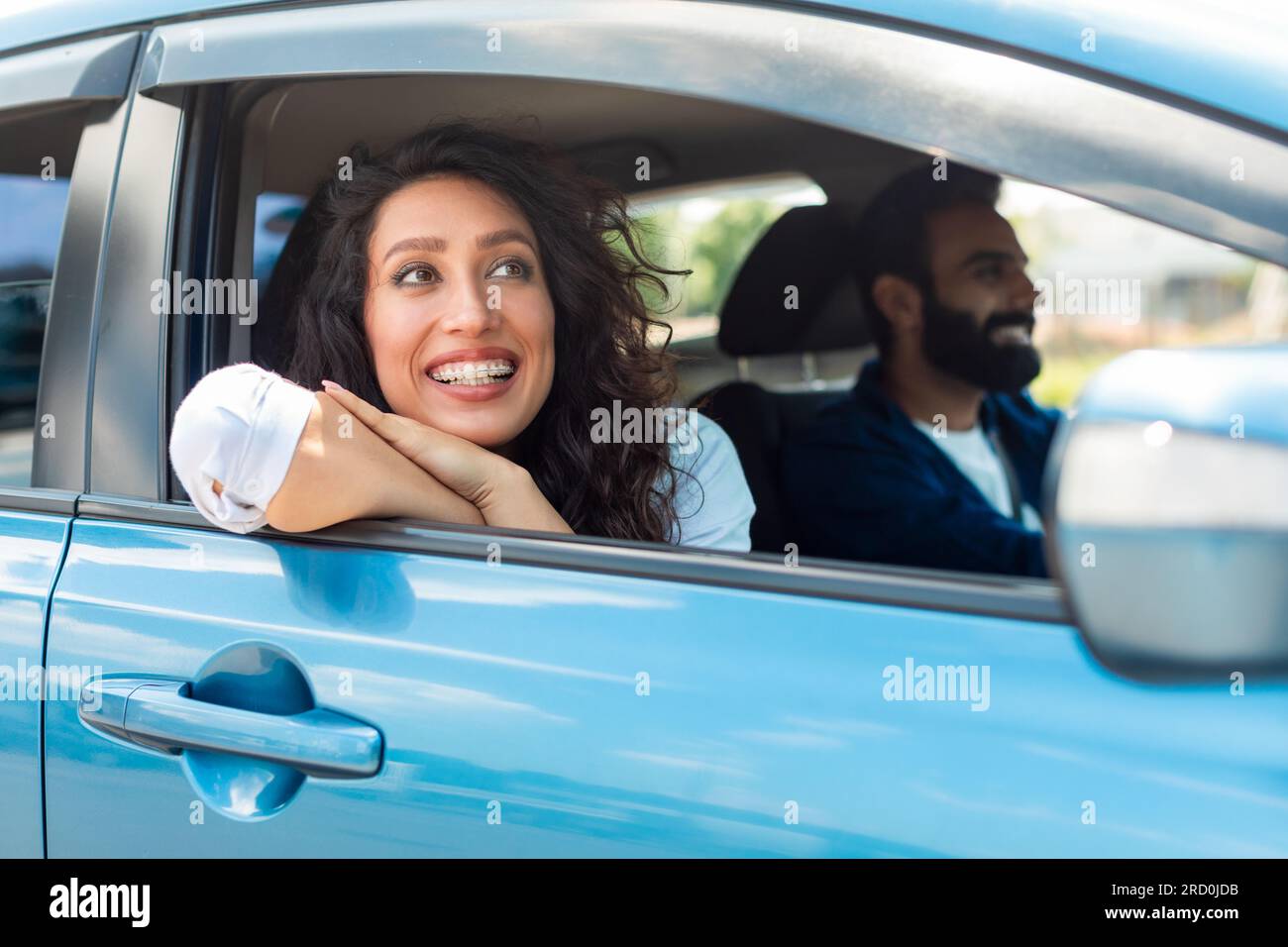 Lächelnde, traumhafte arabische Dame, die im Auto sitzt, aus dem offenen Fenster schaut und lächelt, die Reise genießt, ein Mann, der Auto fährt Stockfoto
