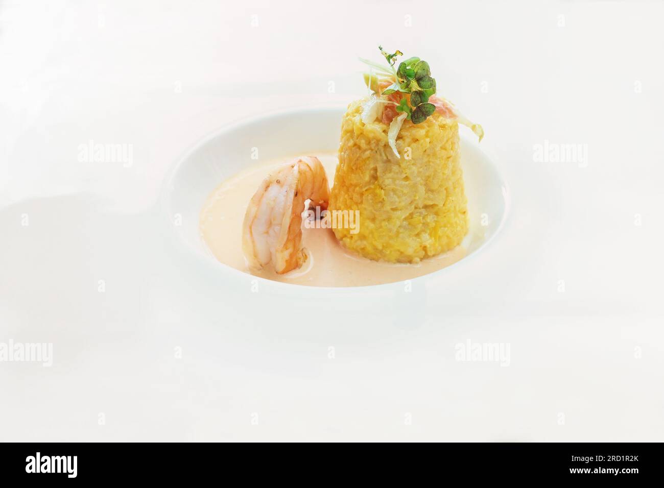 Safran-Risotto in Konusform mit Kräuter- und Gemüsegarnierung, einer großen Garnelen- und Krustentiersauce auf einem modernen weißen Teller, köstliches Fisch- und Meeresfrüchtegericht, C. Stockfoto
