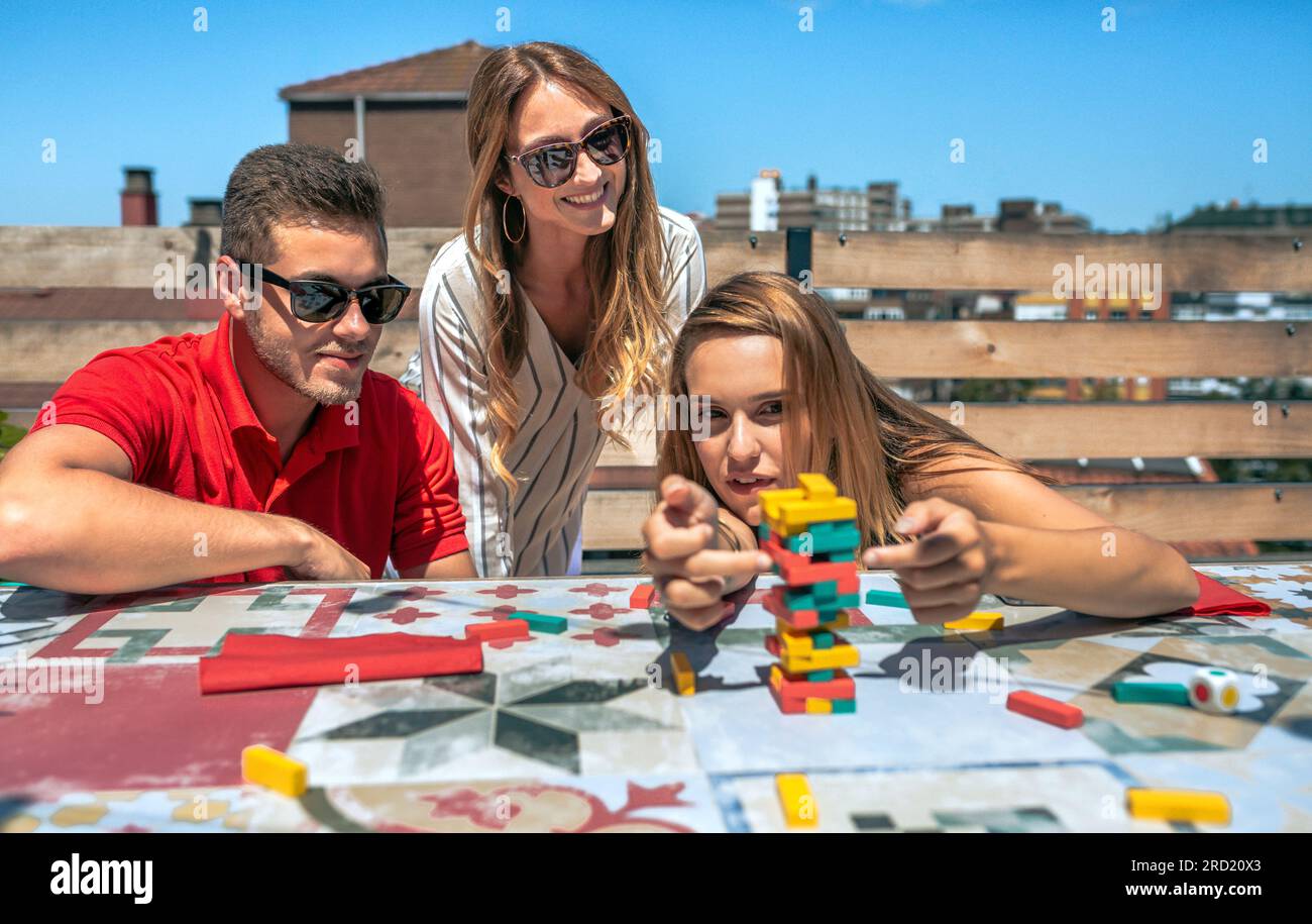 Konzentrierte junge Frau, die auf einer Sommerparty neben ihren lächelnden Freunden Jenga-Spielfigur auf dem Dach schiebt Stockfoto