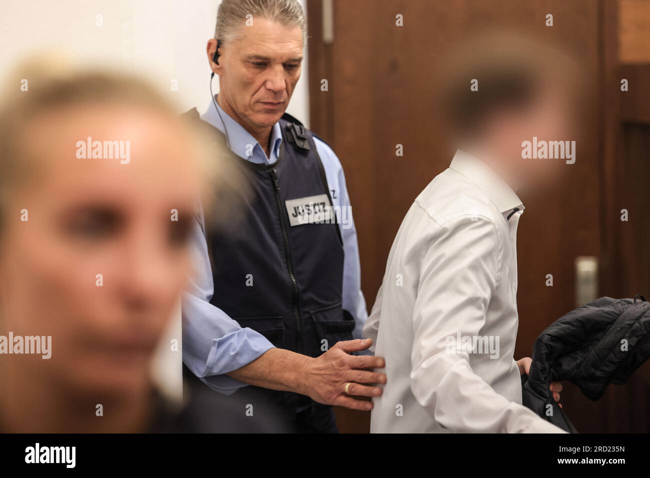 Köln, Deutschland. 18. Juli 2023. Der Angeklagte (r) wird von Gerichtsbediensteten in den Gerichtssaal geführt. Der 22-Jährige soll das Haus seines Großvaters in Gummersbach angezündet haben, um ihn zu töten. Der Mann starb an Rauchvergiftung. Kredit: Oliver Berg/dpa - ACHTUNG: Beklagter auf Anordnung des Gerichts/dpa/Alamy Live News verpixelt Stockfoto