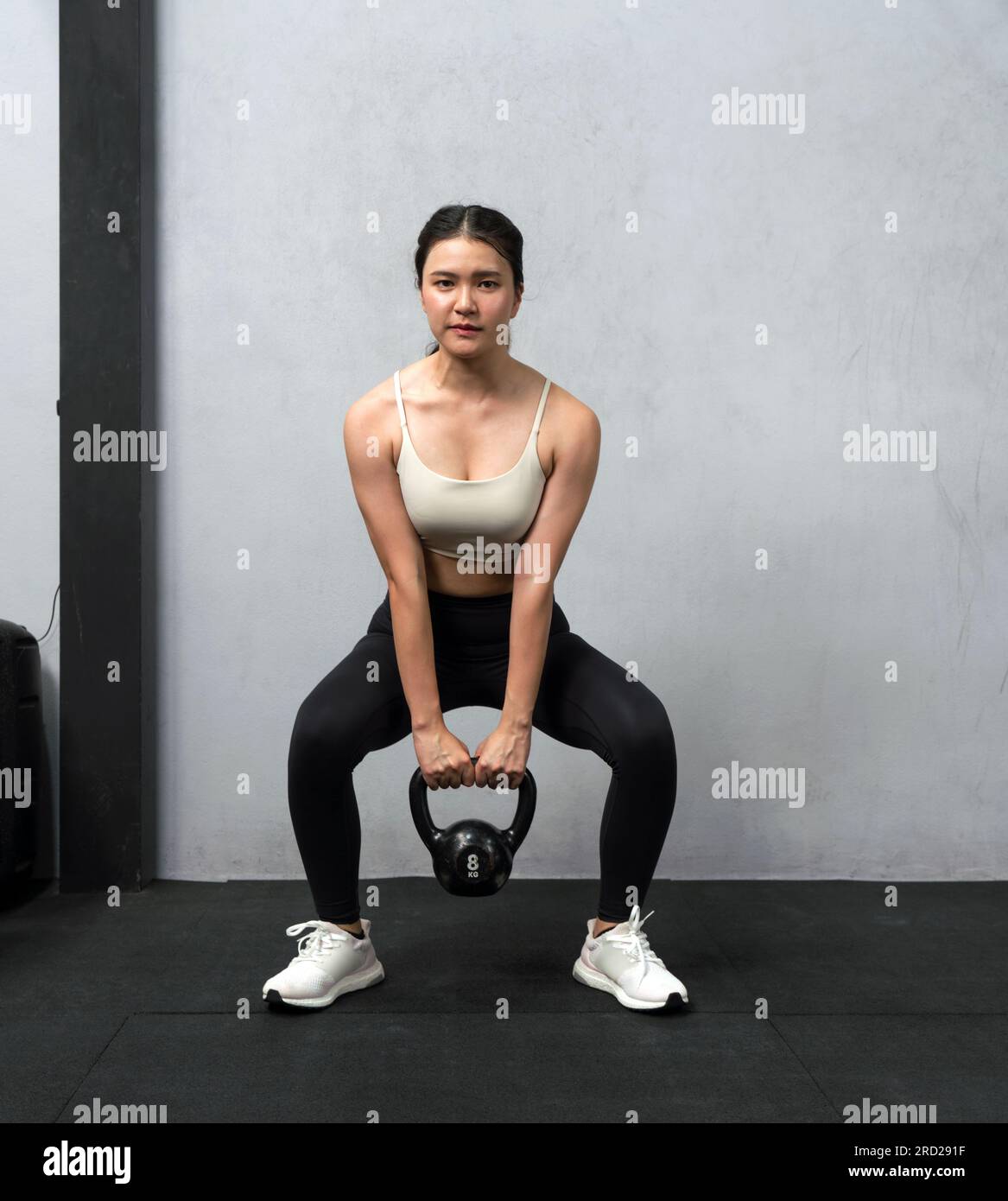 Die junge asiatische Frau tritt in einem gut ausgestatteten Fitnessraum mit Kugelhantel auf. Ihre sportliche Form ist der Inbegriff ausgewogener Kraft und Ausdauer. Sie greift sich einen Krankenwagen an Stockfoto