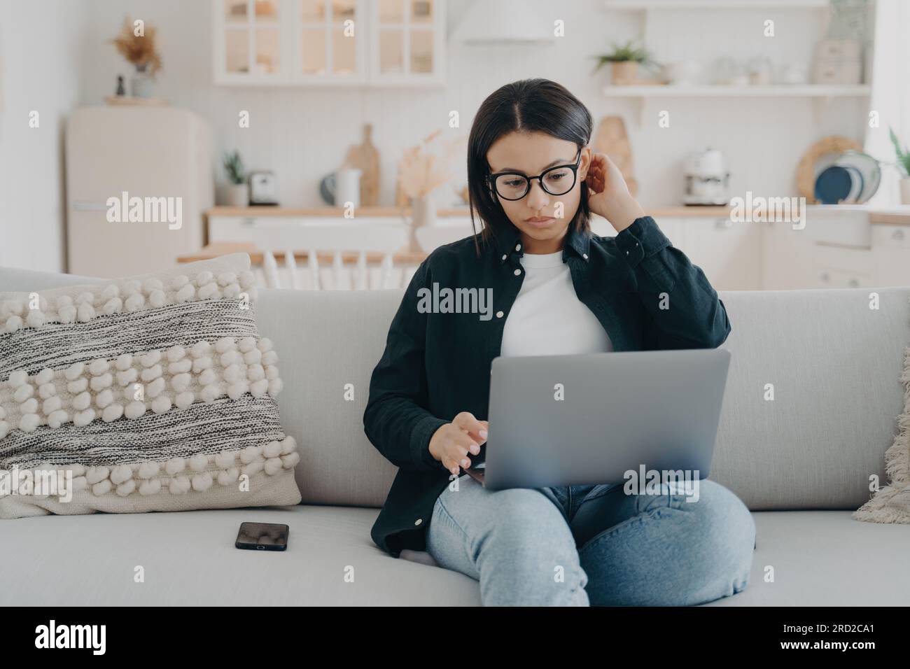 Fokussierte Freiberuflerin mit Brille, die auf einem Laptop arbeitet und zu Hause auf dem Sofa sitzt. Konzentrierte junge Frauen checken E-Mails, soziale Netzwerke, Einkaufen Stockfoto