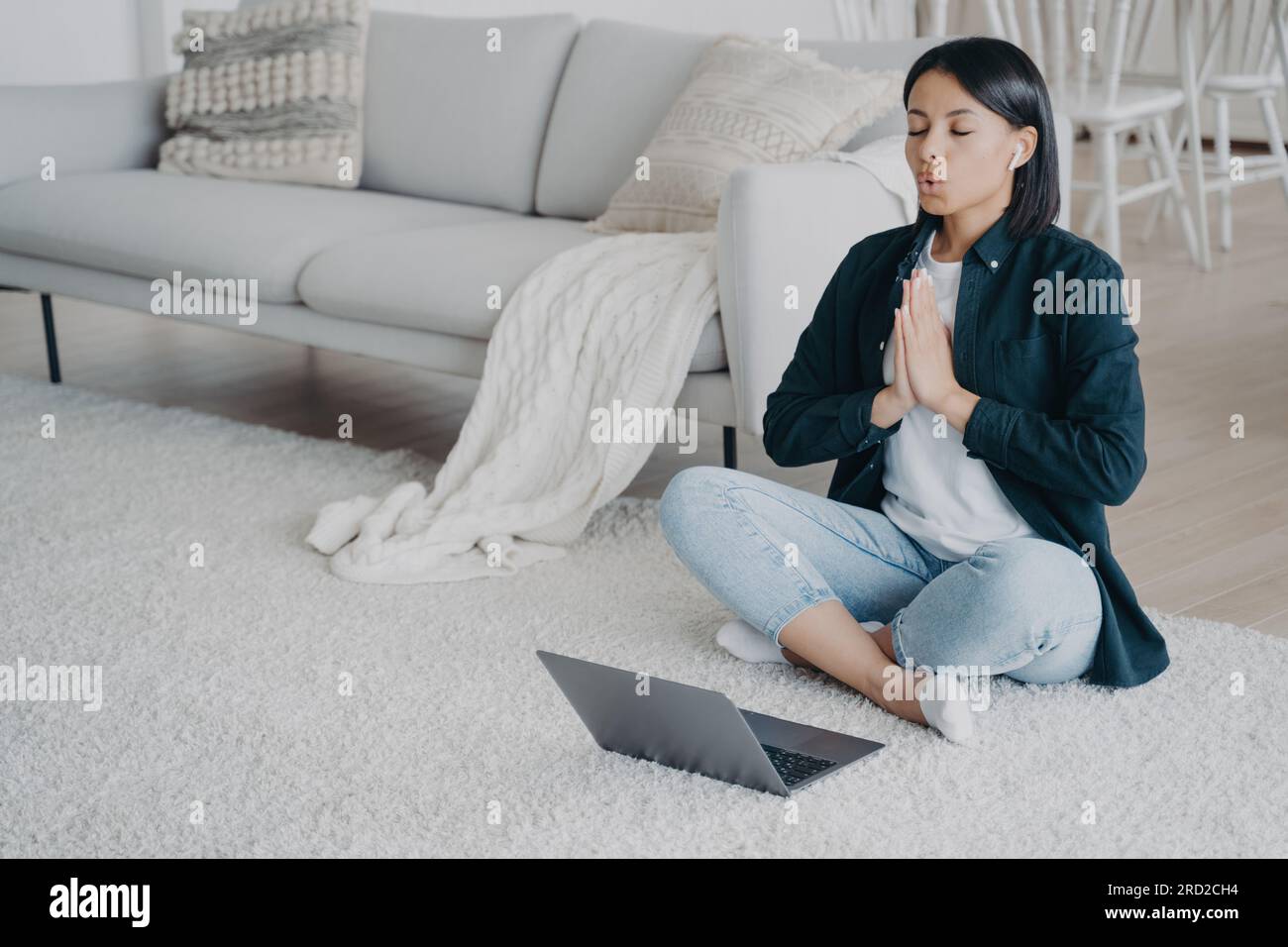 Junge europäische Frau, die zu Hause online Yoga praktiziert. Ein Mädchen, das auf dem Boden vor dem Laptop sitzt, in Lotusstellung und meditiert. Musik hören, ruhig sein Stockfoto