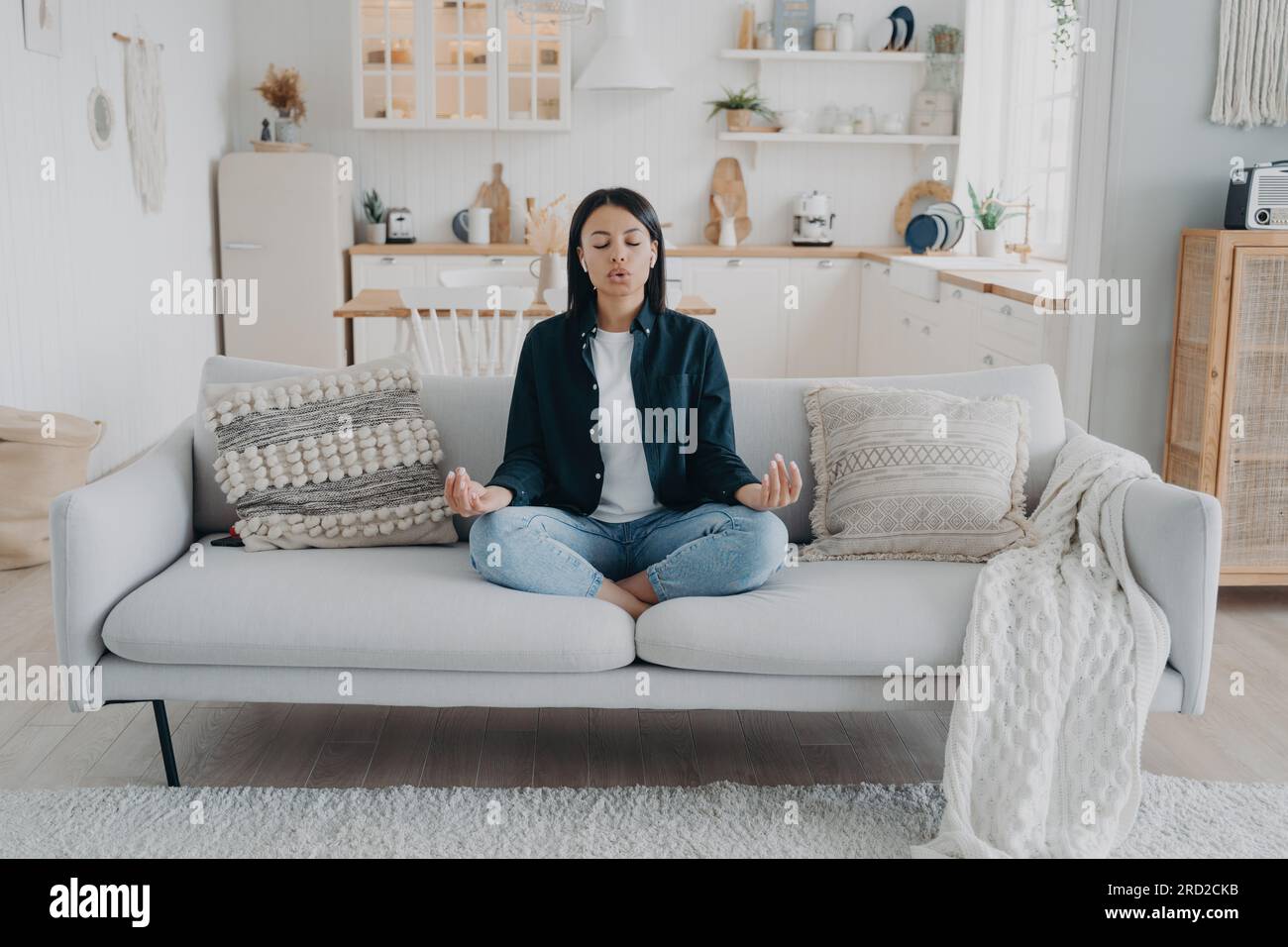 Eine ruhige Frau übt Yoga mit Mudra-Geste, atmet auf dem Sofa im Wohnzimmer. Junge Frau meditiert Stress lindern, zu Hause entspannen. Heilung Stockfoto