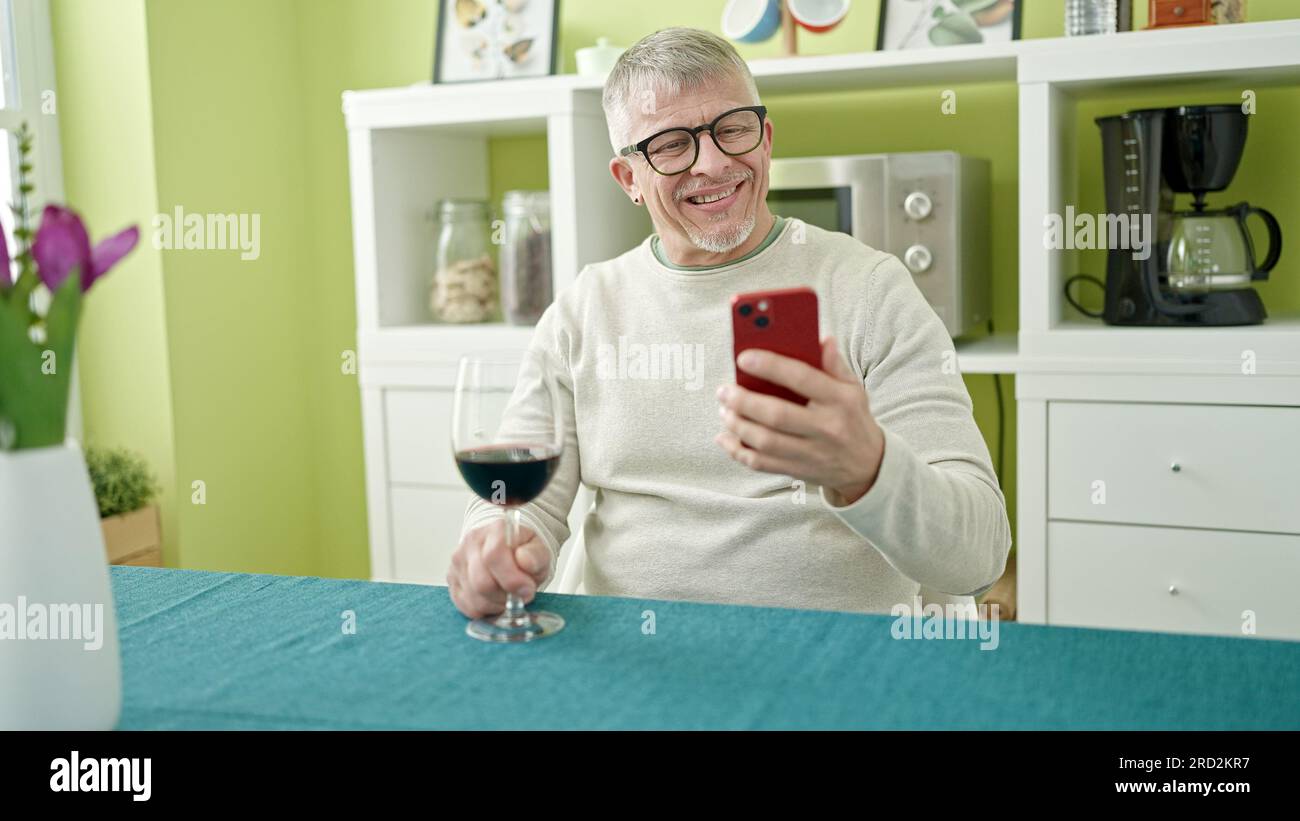 Ein grauhaariger Mann im mittleren Alter, der zu Hause ein Glas Wein mit Smartphone trinkt Stockfoto