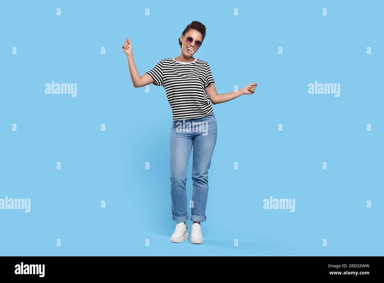 Glückliche junge Frau in stilvoller Sonnenbrille, die auf hellblauem Hintergrund tanzt Stockfoto