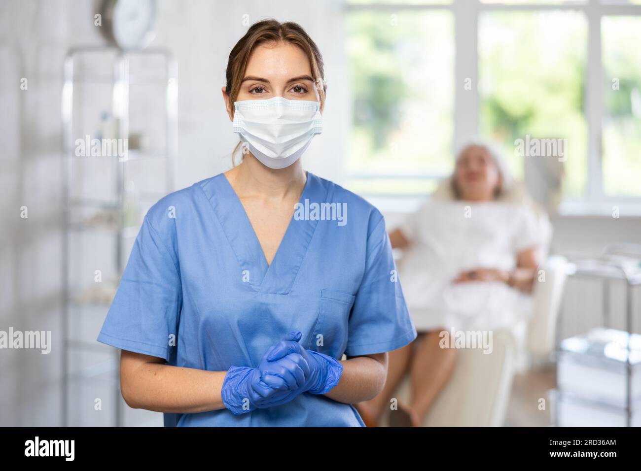 Glücklicher Arzt oder Krankenschwester mit Maske in Uniform mit gekreuztem Arm, Patient hinter dem Sofa. Stockfoto