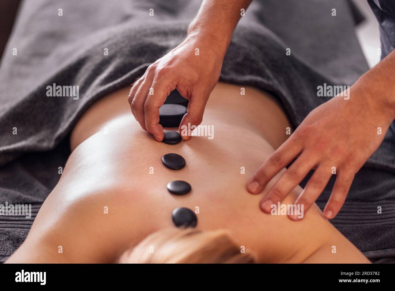 Nahaufnahme der männlichen Hände eines Masseurs, der eine Massage mit heißen Steinen an seine Klientin macht. Ein großer schwarzer Kiesel auf dem weiblichen Rücken, entlang der Wirbelsäule. Stockfoto