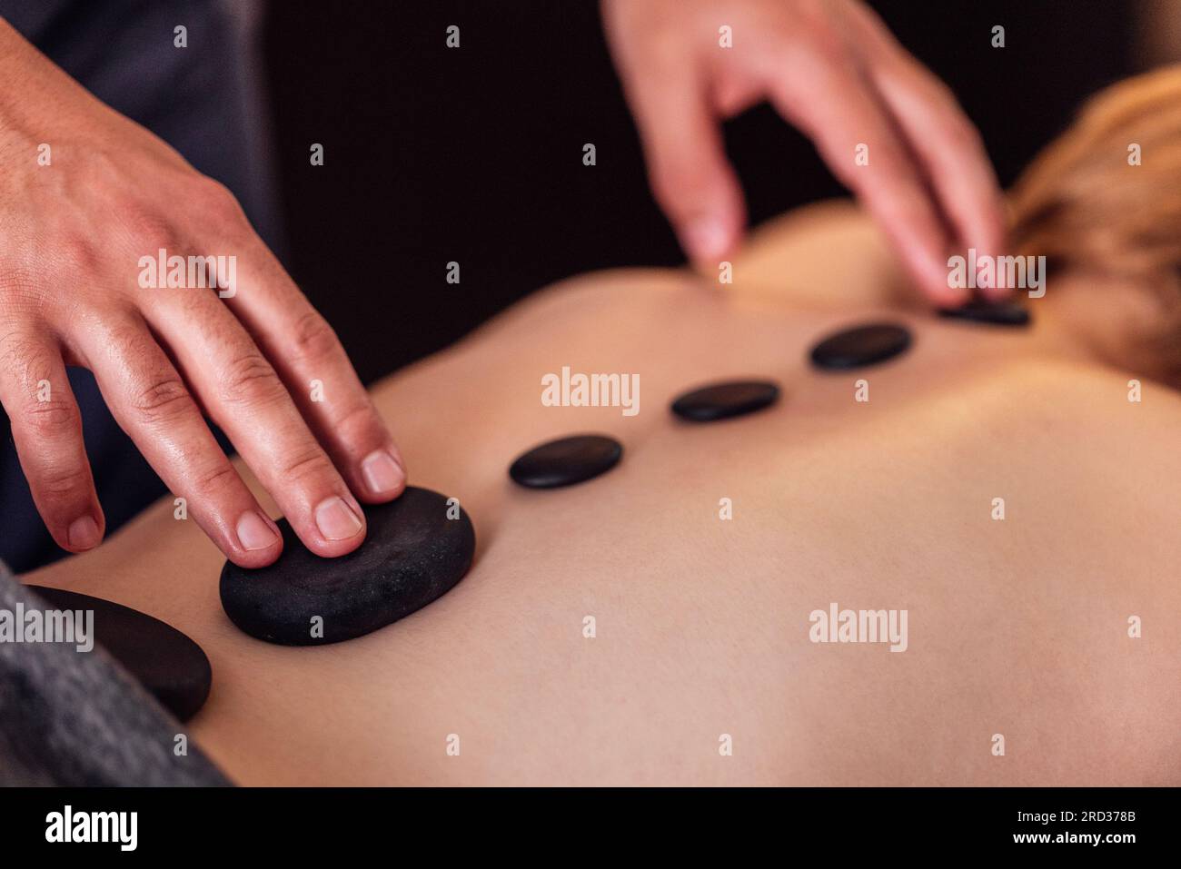 Nahaufnahme der männlichen Hände eines Masseurs, der eine Massage mit heißen Steinen an seine Klientin macht. Ein großer schwarzer Kiesel auf dem weiblichen Rücken, entlang der Wirbelsäule. Stockfoto