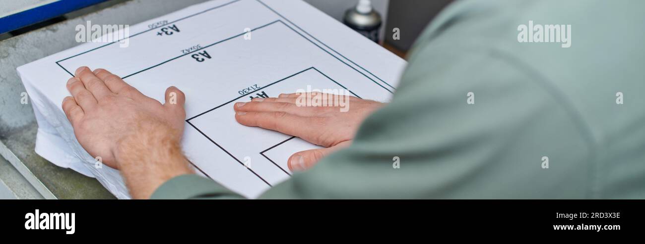 Zugeschnittene Ansicht eines jungen Handwerkers, der ein T-Shirt in der Nähe einer Siebdruckmaschine im Druckstudio hält, kundenorientiertes Konzept für kleine Unternehmen, cr Stockfoto