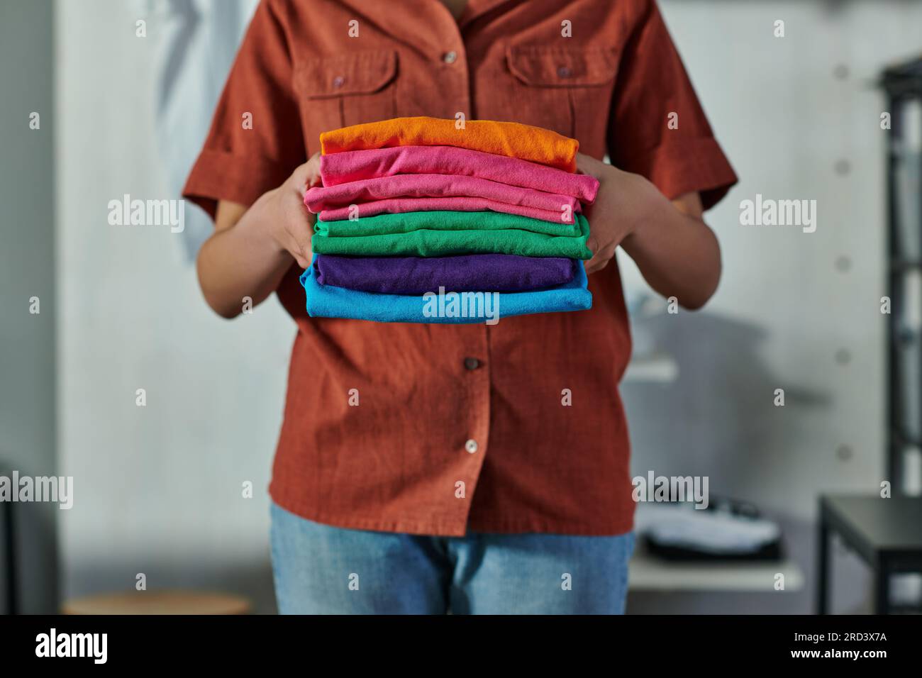 Zugeschnittener Blick auf eine verschwommene junge afroamerikanische Kunsthandwerkerin, die Kleidung hält, während sie in einem verschwommenen Druckstudio im Hintergrund arbeitet und steht, Kunden- Stockfoto