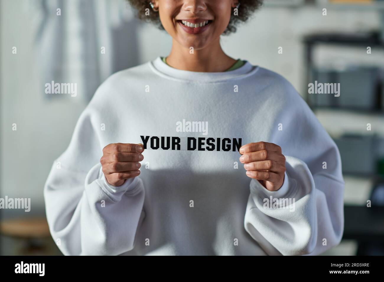 Freigestellte Ansicht einer lächelnden jungen afroamerikanischen Kunsthandwerkerin, die die Druckschicht mit Ihrem Designtext hält und im Print Studio, fokussierten Bus steht Stockfoto