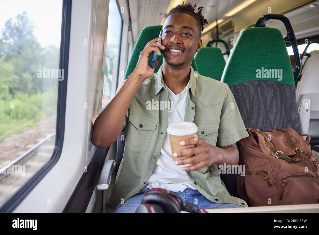 Junger Mann, Der Im Zug Zur Arbeit Pendelt, Im Zug Sitzt Und Mit Einem Handy Mit Take-Away-Kaffee Spricht Stockfoto