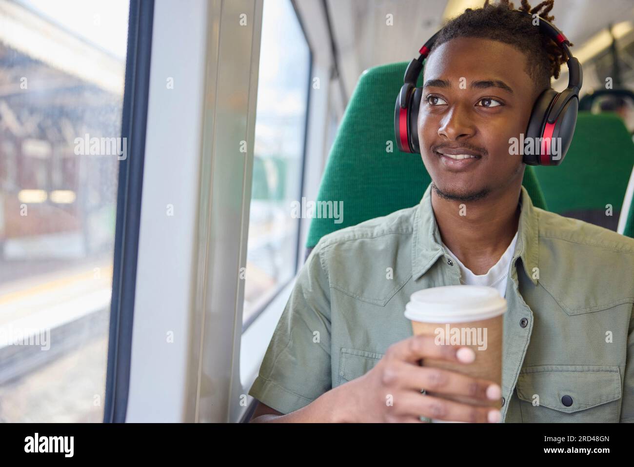 Junger Mann, Der Zur Arbeit Pendelt, Sitzt Im Zug Und Trägt Kabellose Kopfhörer Mit Take-Away-Kaffee Stockfoto