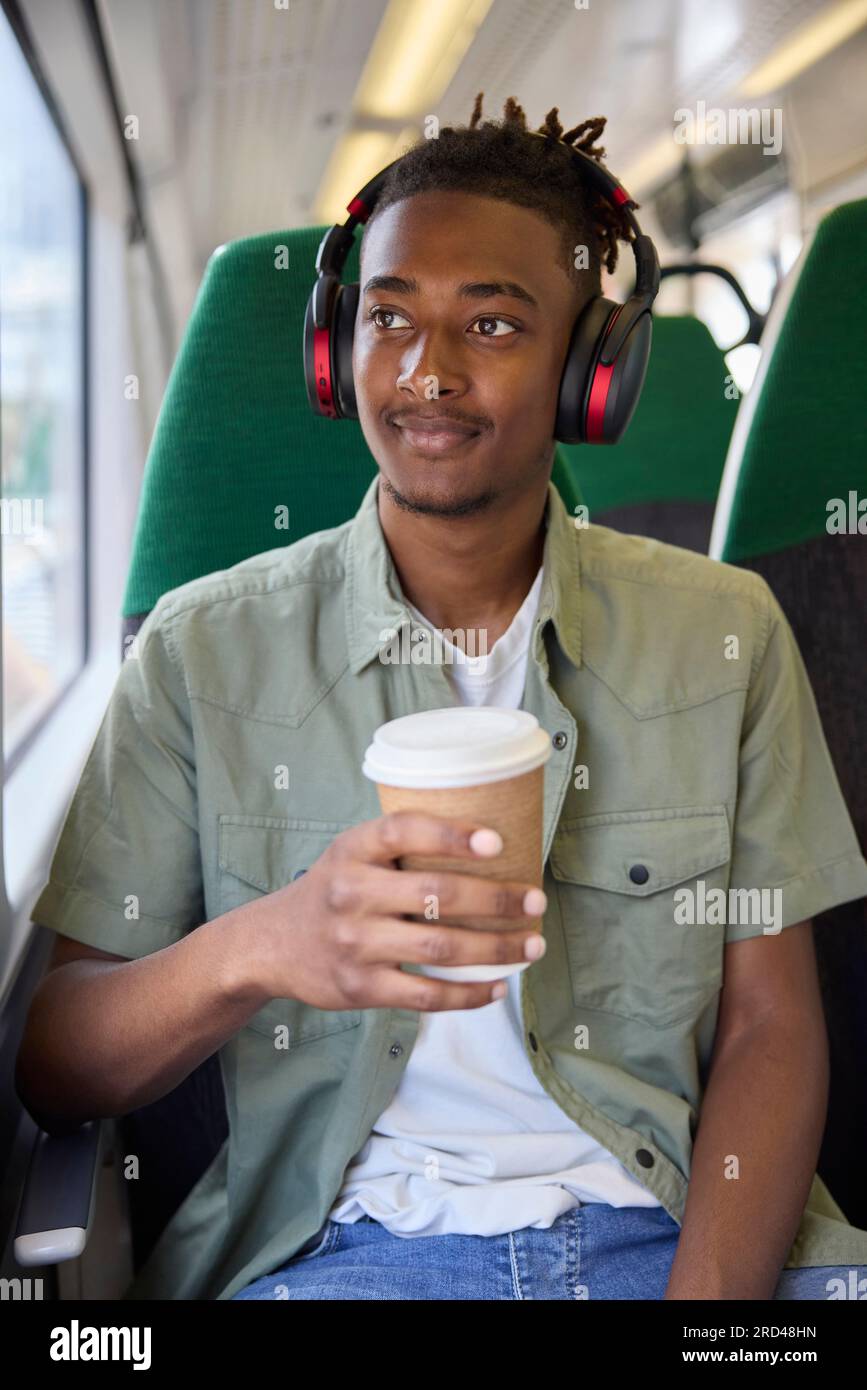 Junger Mann, Der Zur Arbeit Pendelt, Sitzt Im Zug Und Trägt Kabellose Kopfhörer Mit Take-Away-Kaffee Stockfoto