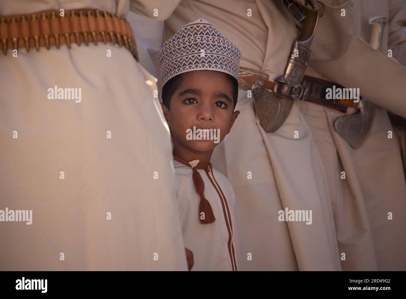 Ein Junge in einer traditionellen omanischen Dishdasha und einem Zanzibari-Hut zieht sich aus einer Menge erwachsener Männer hervor, während sie einen Tanz vorführen. Stockfoto