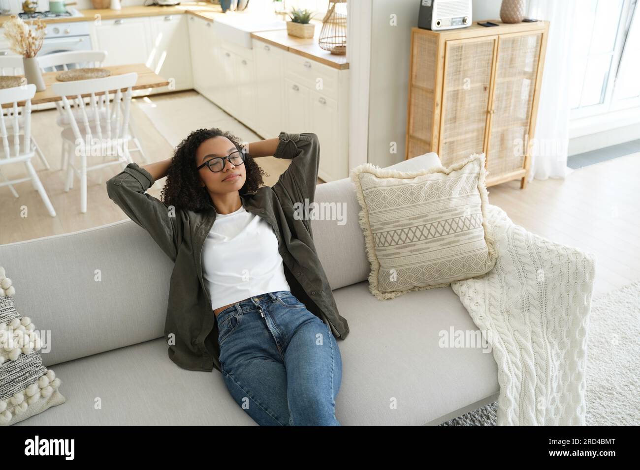 Ruhige afroamerikanische Frau auf einer gemütlichen Couch, Hände hinter dem Kopf, genießt ein faules Wochenende zu Hause. Entspannt, erholsam in komfortabler Umgebung mit frischem Stockfoto