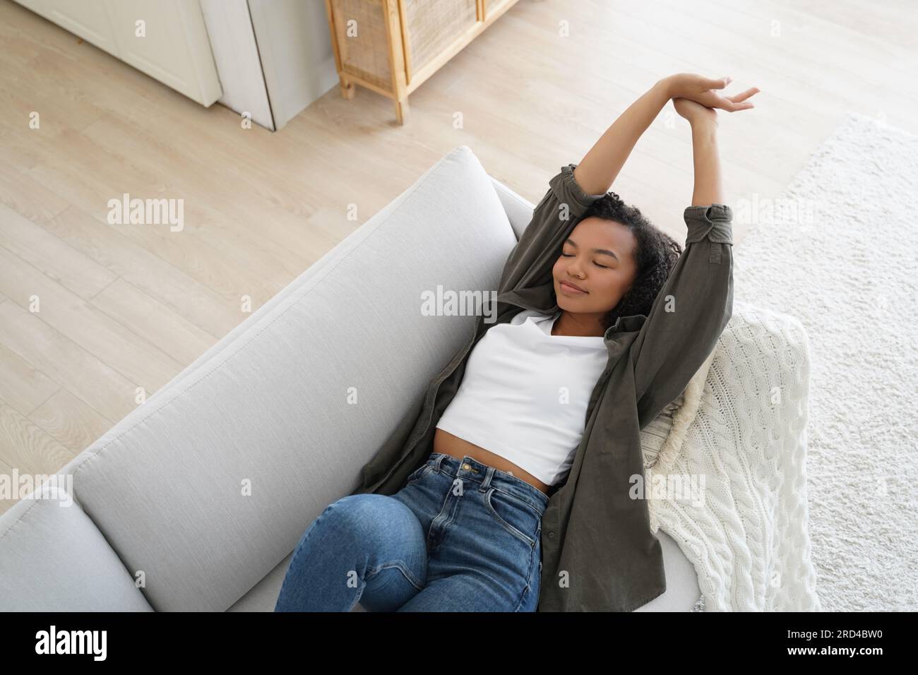 Ruhiges afroamerikanisches Mädchen, das zu Hause auf einem bequemen Sofa schläft und eine entspannende Pause an einem faulen Wochenende genießt. Entspannte, junge Frau, die Arme streckt, Stockfoto