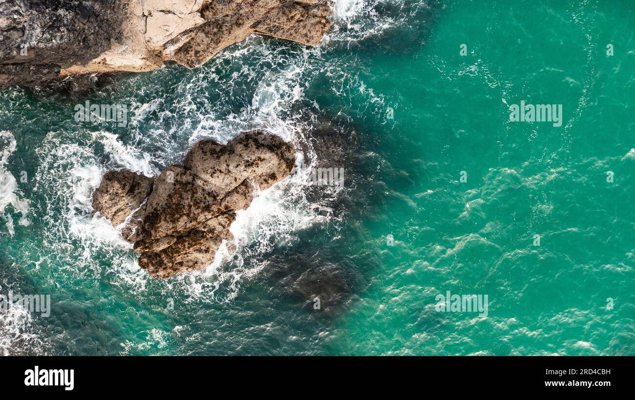 Blick aus der Vogelperspektive direkt über weißen Wellen, die auf rauen Felsen brechen, in einer rauen Umgebung an der Küste von Cornwall mit Platz zum Kopieren über Smaragdgrün Stockfoto