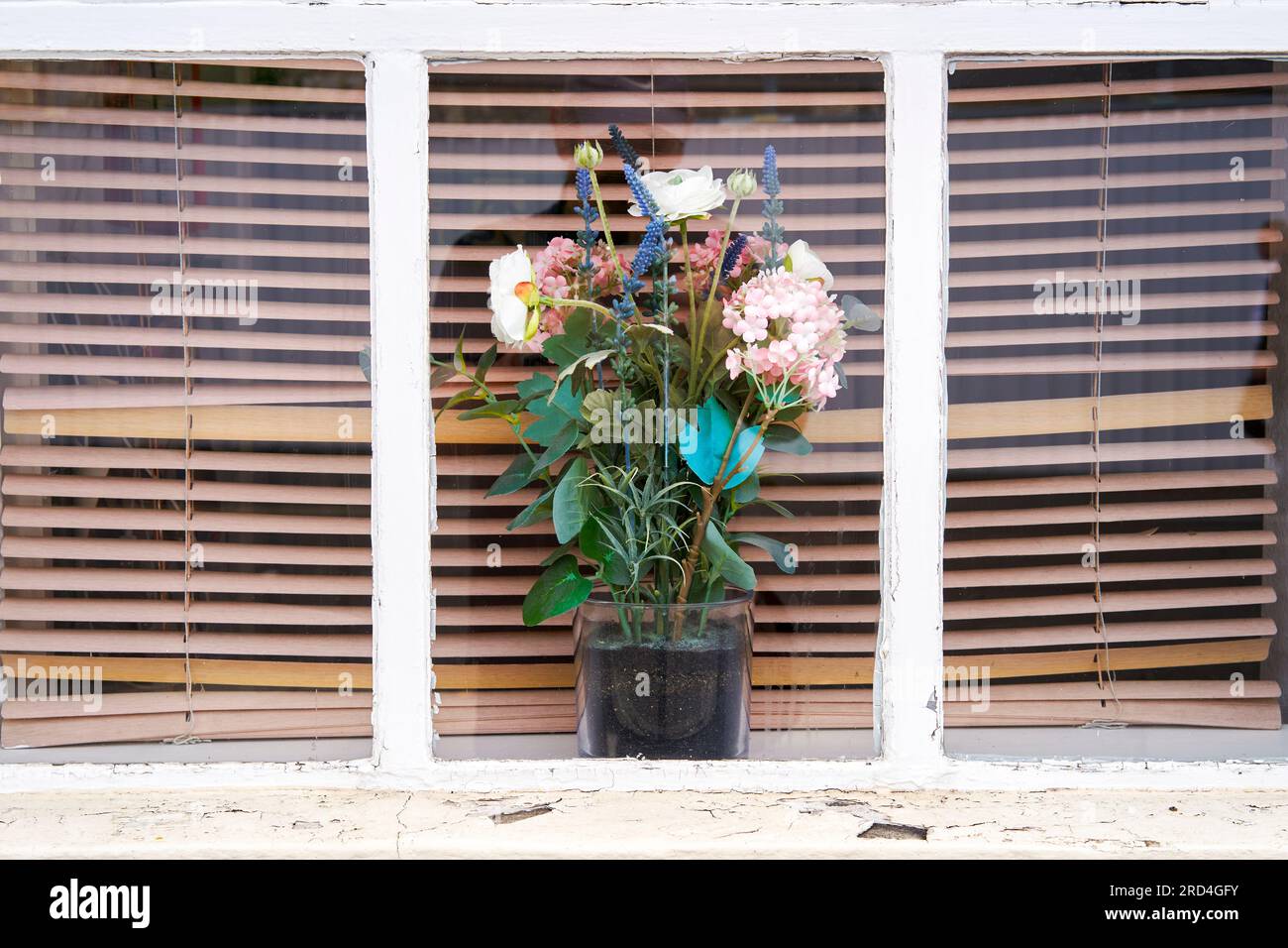 Plastikblumen in einem Topf in einem Fenster mit Jalousien dahinter Stockfoto